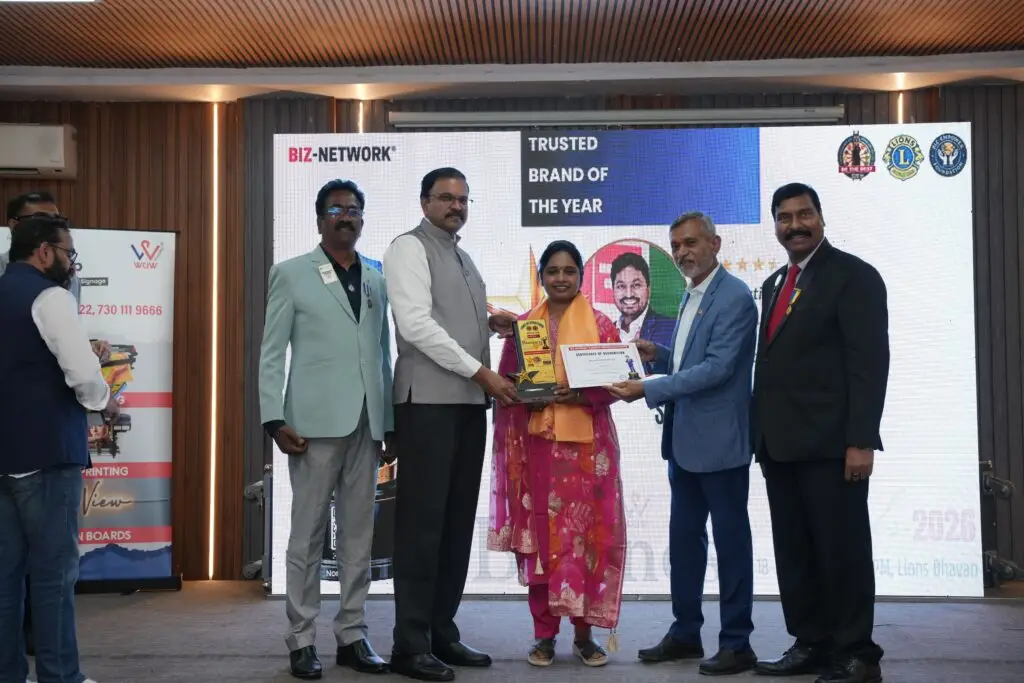 Dr. Akhila Reddy receiving the "Trusted Brand of the Year" award on stage from Lions International officials. She is holding both a trophy and a certificate of merit in front of a large digital screen displaying the award title and the Biz-Network logo.